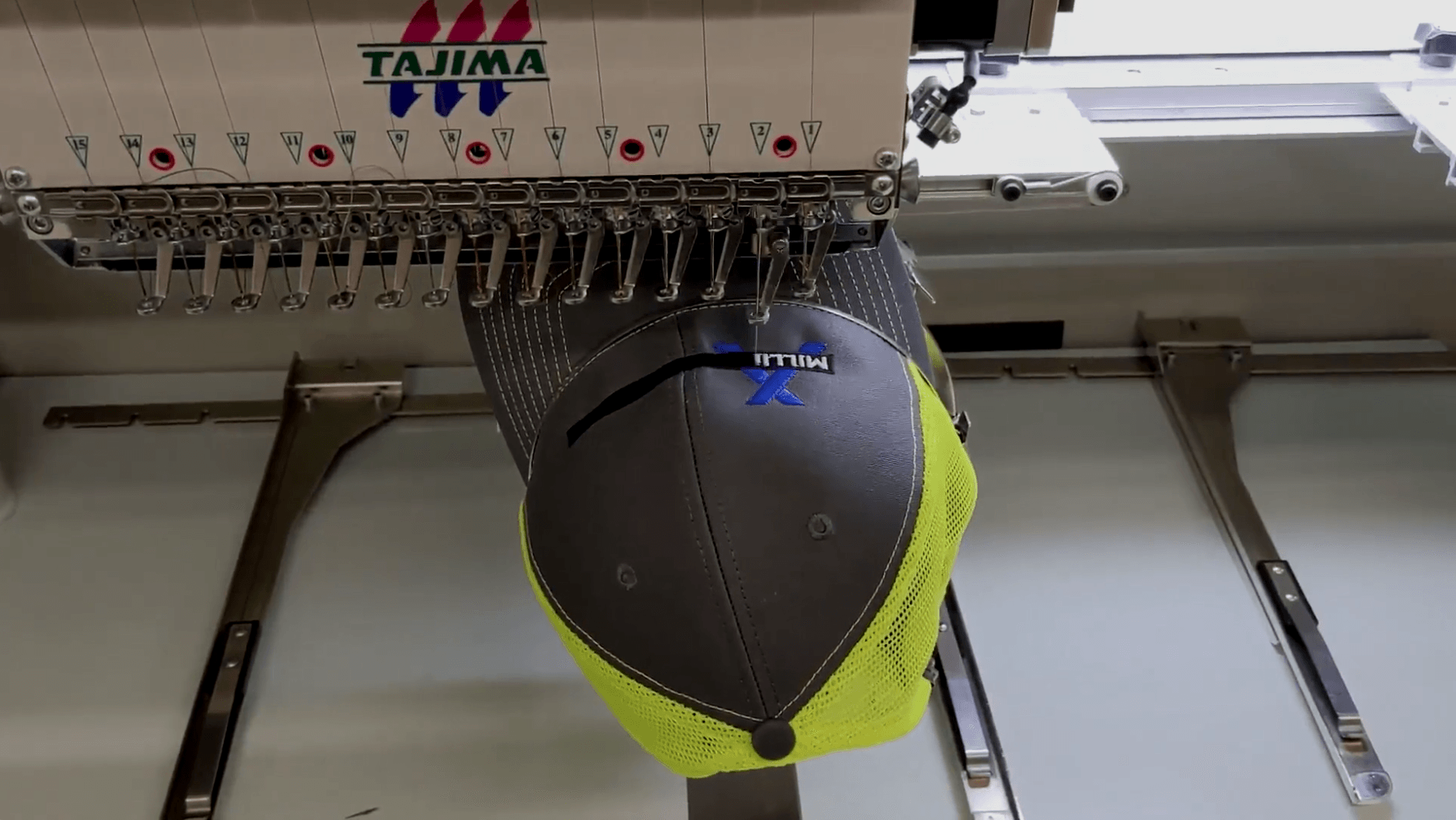 Embroidery machine stitching a gray and neon yellow cap with a blue logo.