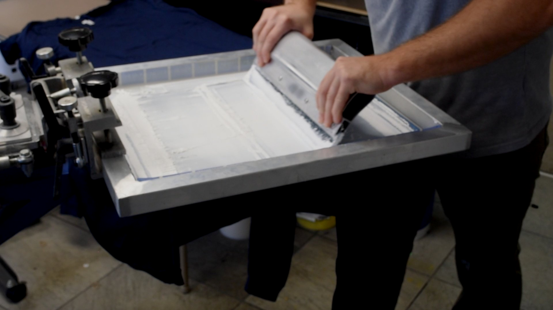 Person pulling a white screen-printing frame across a press in a workshop