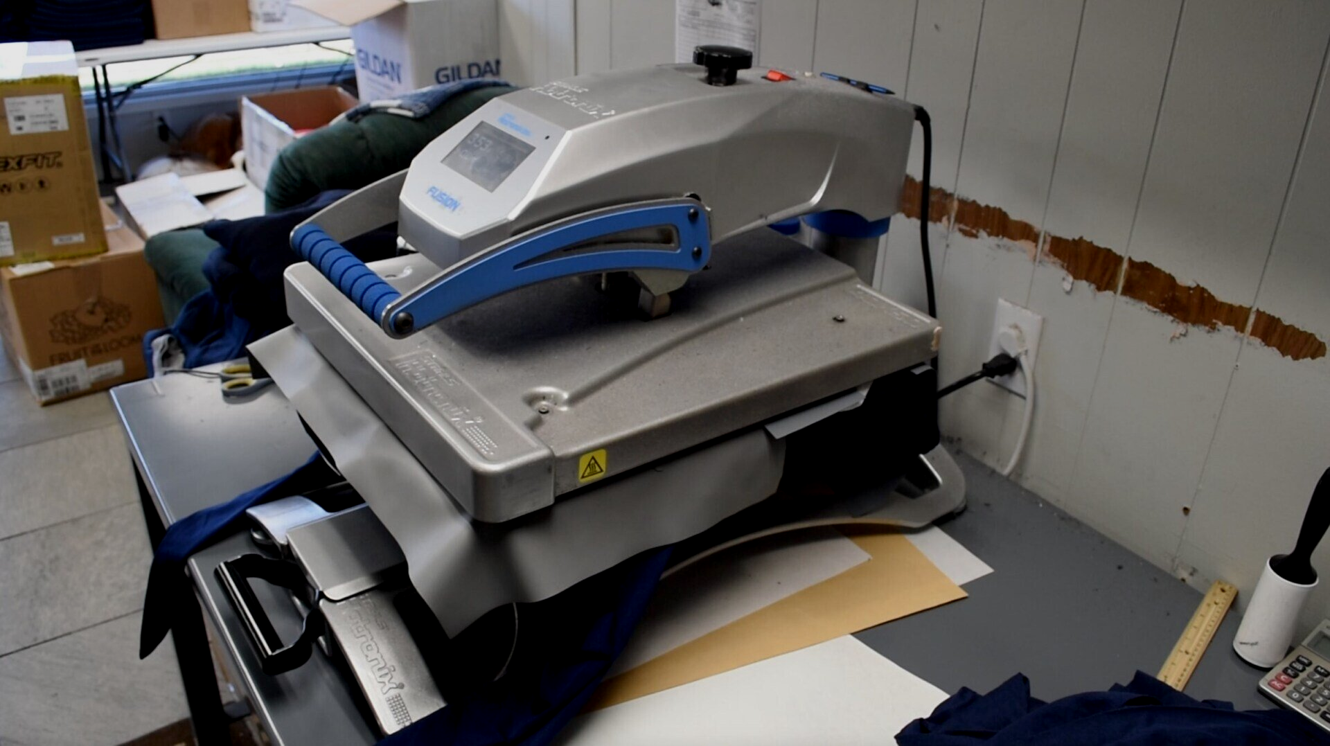 Large gray heat transfer machine on a cluttered workbench with boxes and fabric nearby in a workshop.