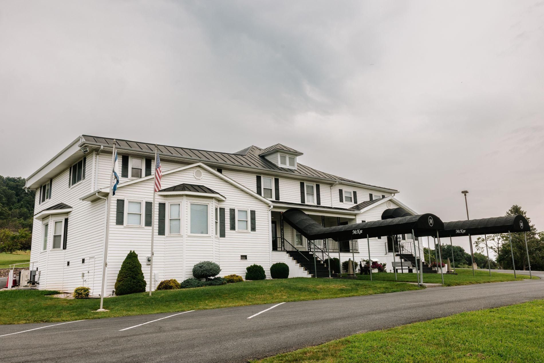White two-story house with dark roof and carport along a roadside under a cloudy sky