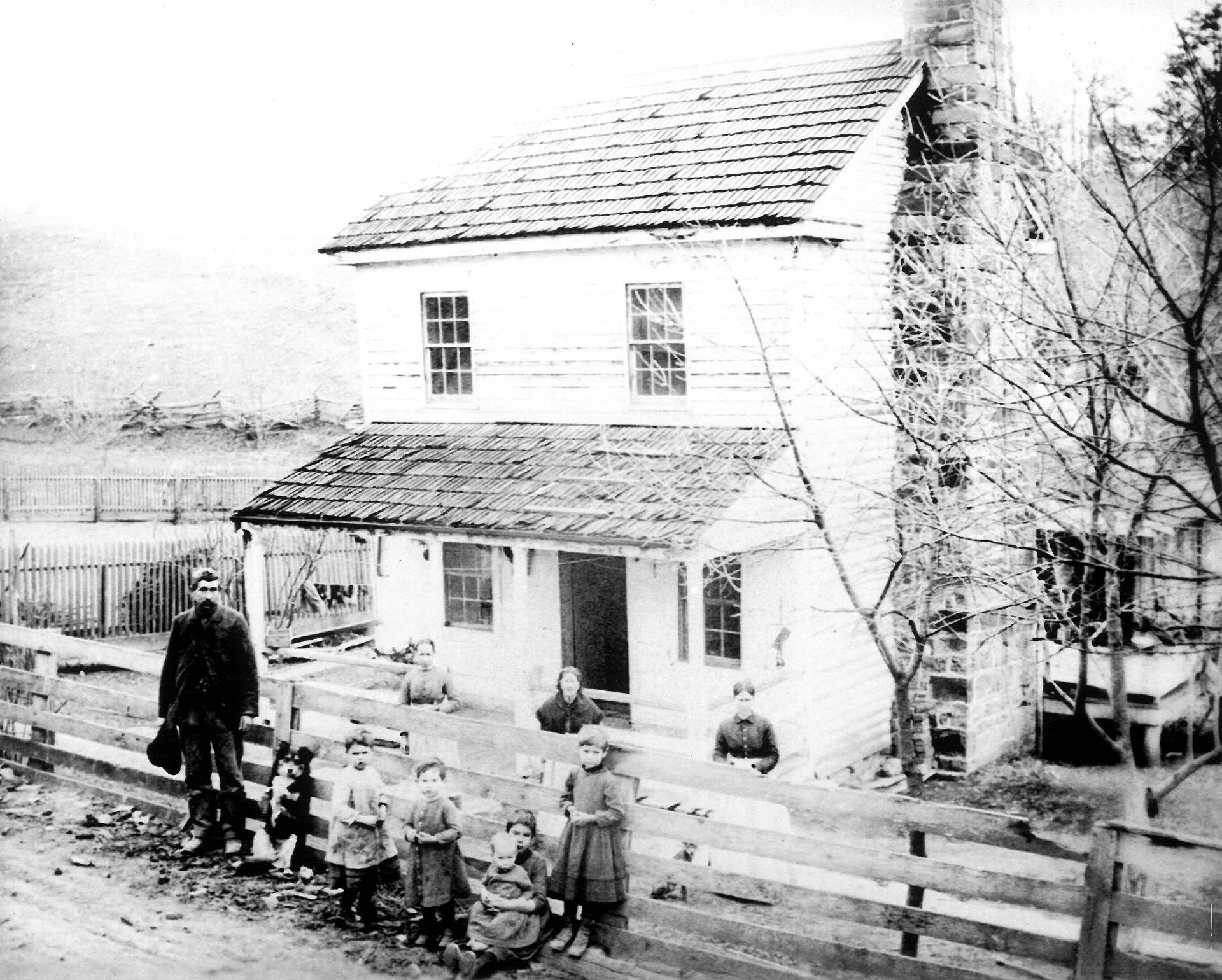 Black-and-white photo of a farmhouse with a porch, people, and a horse near a wooden fence.