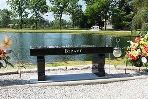 Black memorial bench engraved “Brewer” beside flowers and a pond in a sunny park