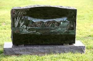 Black granite gravestone with engraved landscape scene on a grassy lawn.