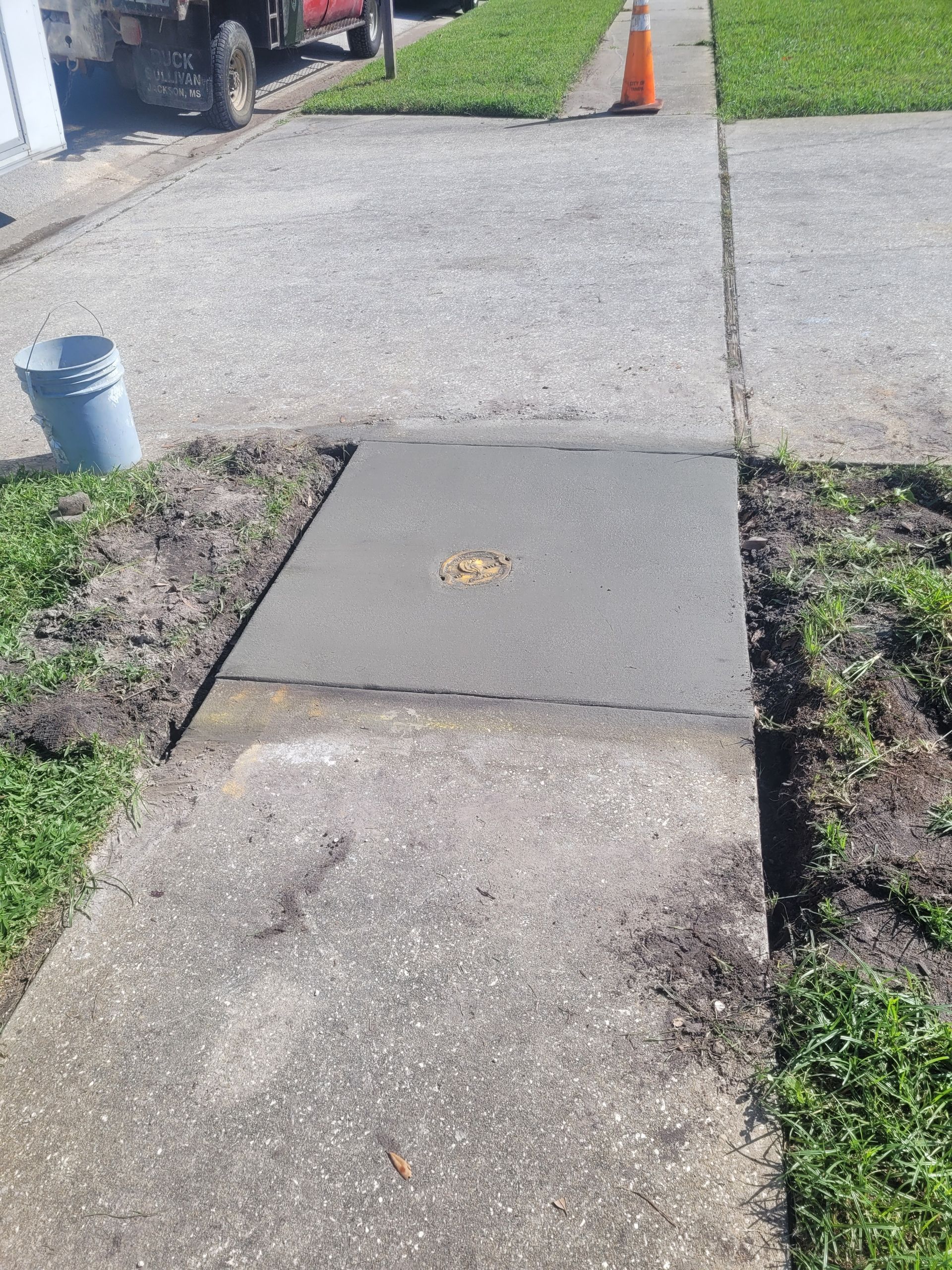 Newly poured concrete square replaces a section of sidewalk, surrounded by dirt and grass, with a bucket and cone nearby.