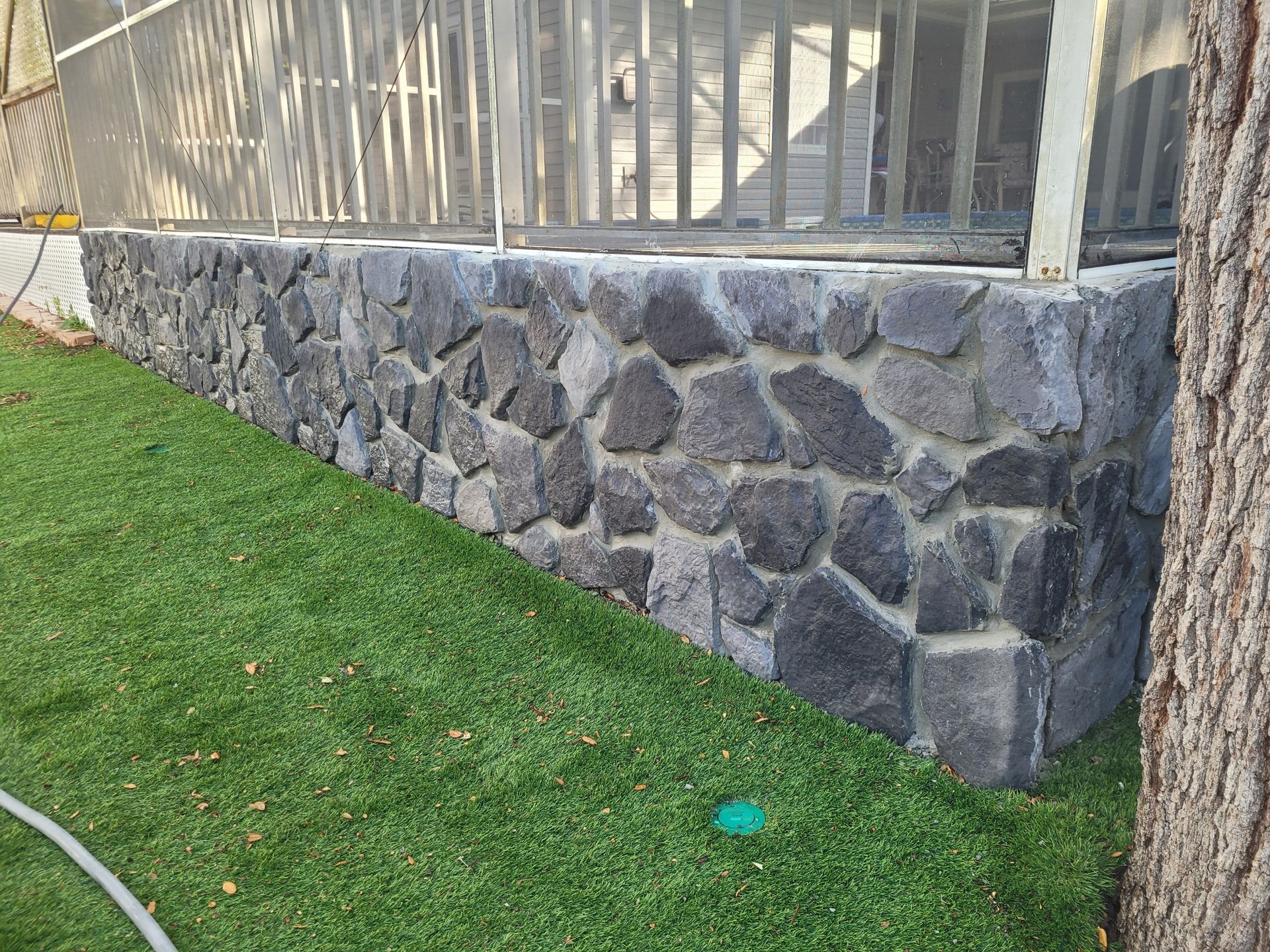 Stone wall supporting a porch. Artificial green lawn in front. Tree on the right.