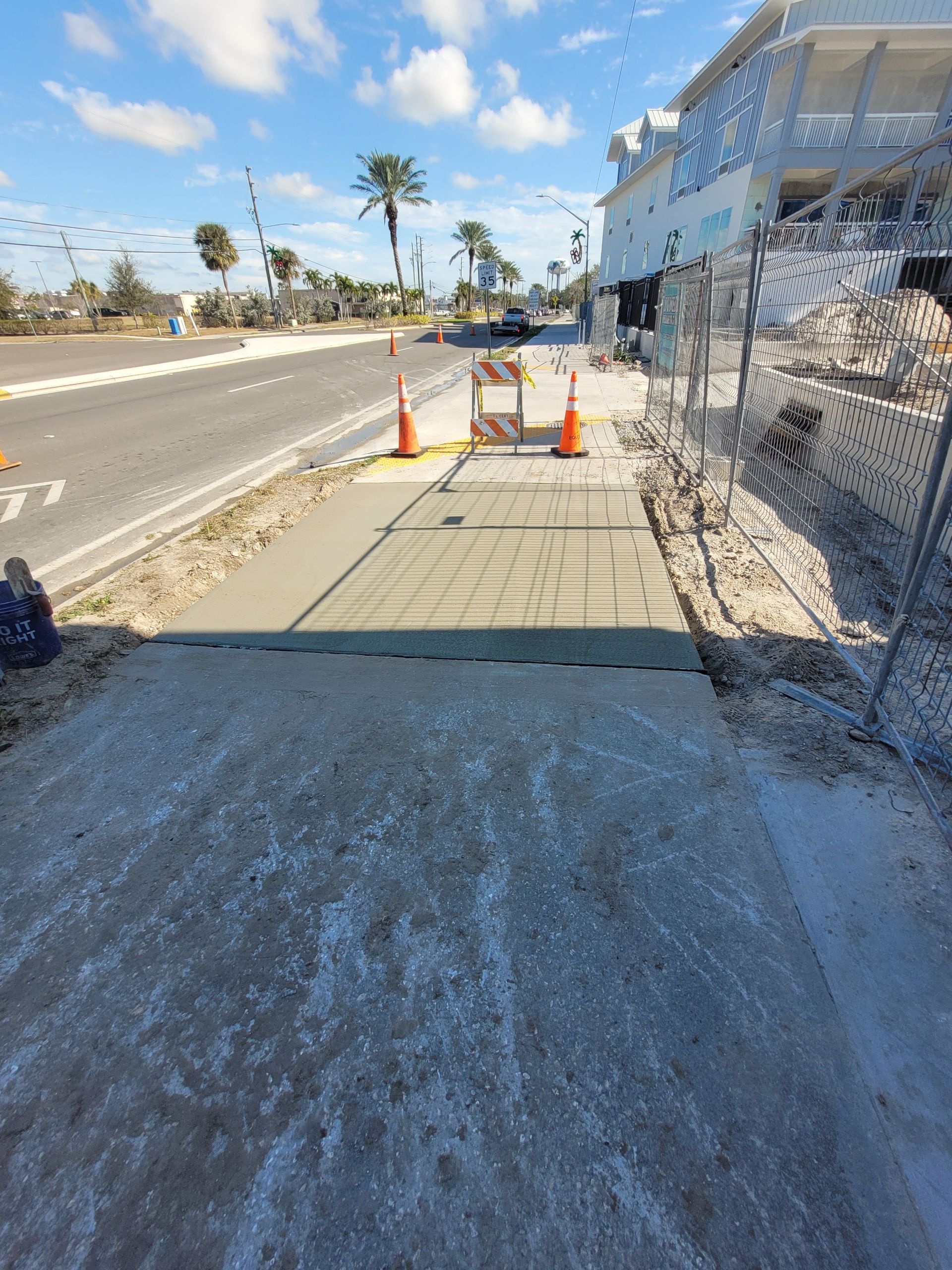 Sidewalk construction with wet concrete and cones blocking access. Building and street in background.