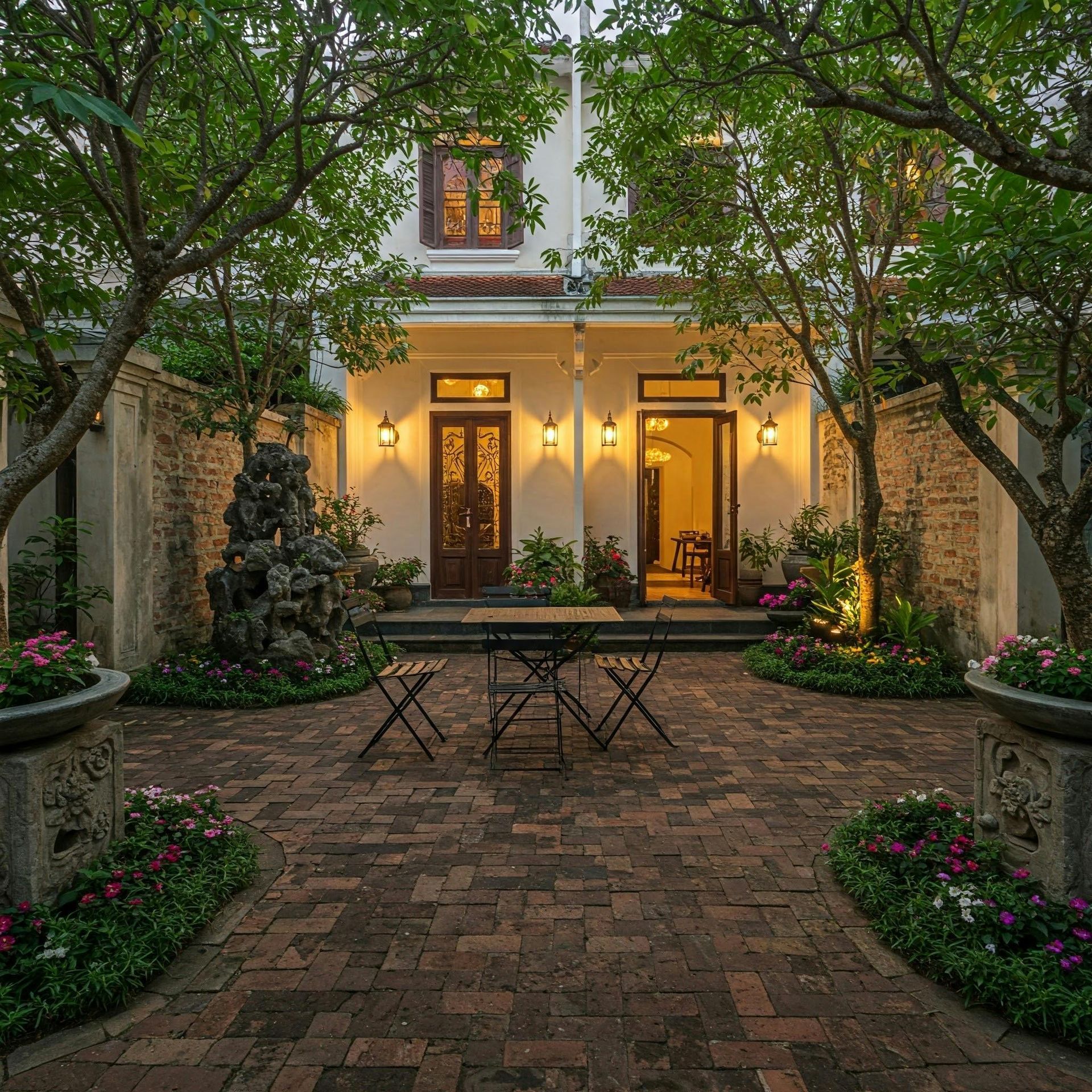 Brick courtyard with table and chairs, surrounded by trees, leading to a house with illuminated doorway.