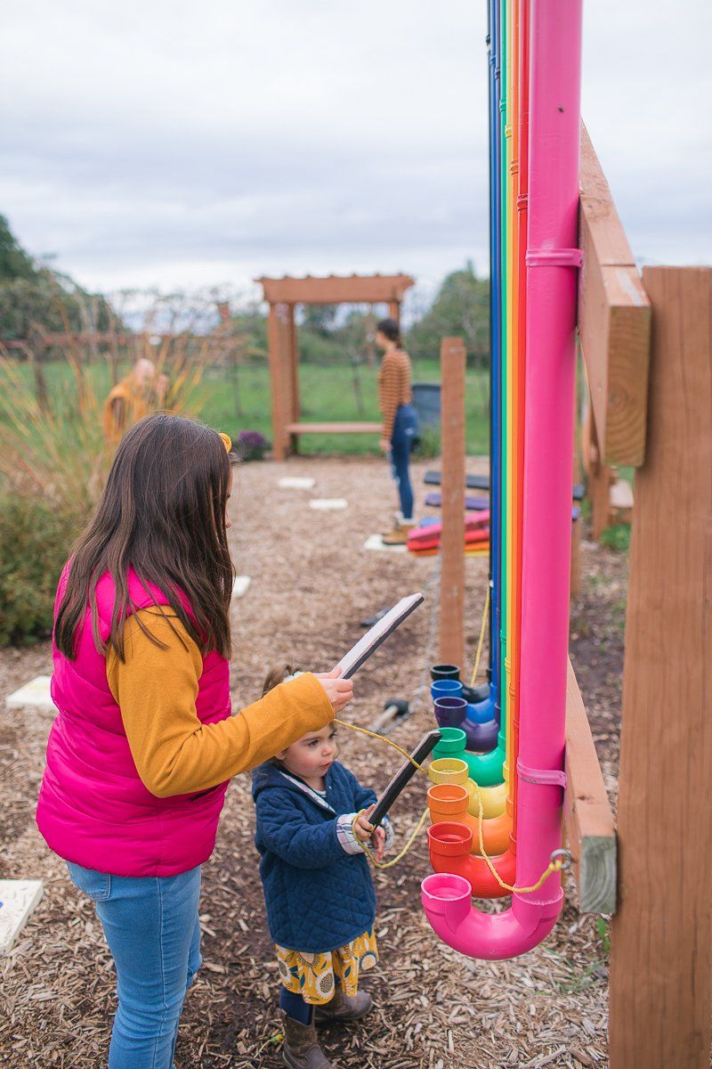 Two children play a rainbow-colored outdoor musical instrument. One child holds a mallet and the other holds a clipboard.