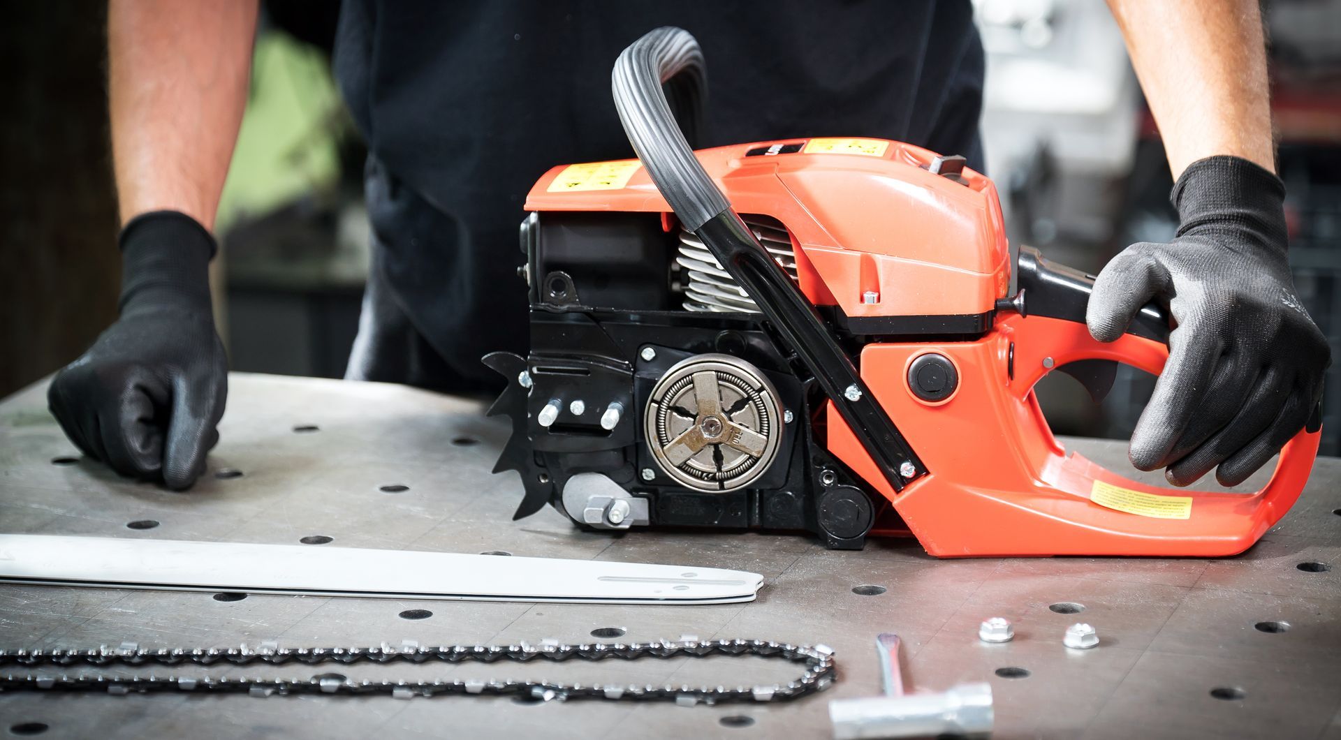 Person in black gloves handling an orange chainsaw on a workbench with a guide bar and chain nearby