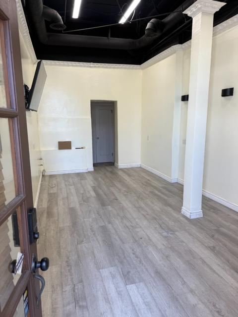 Empty room with white walls, gray wood floor, tall columns, and a doorway under a black ceiling.
