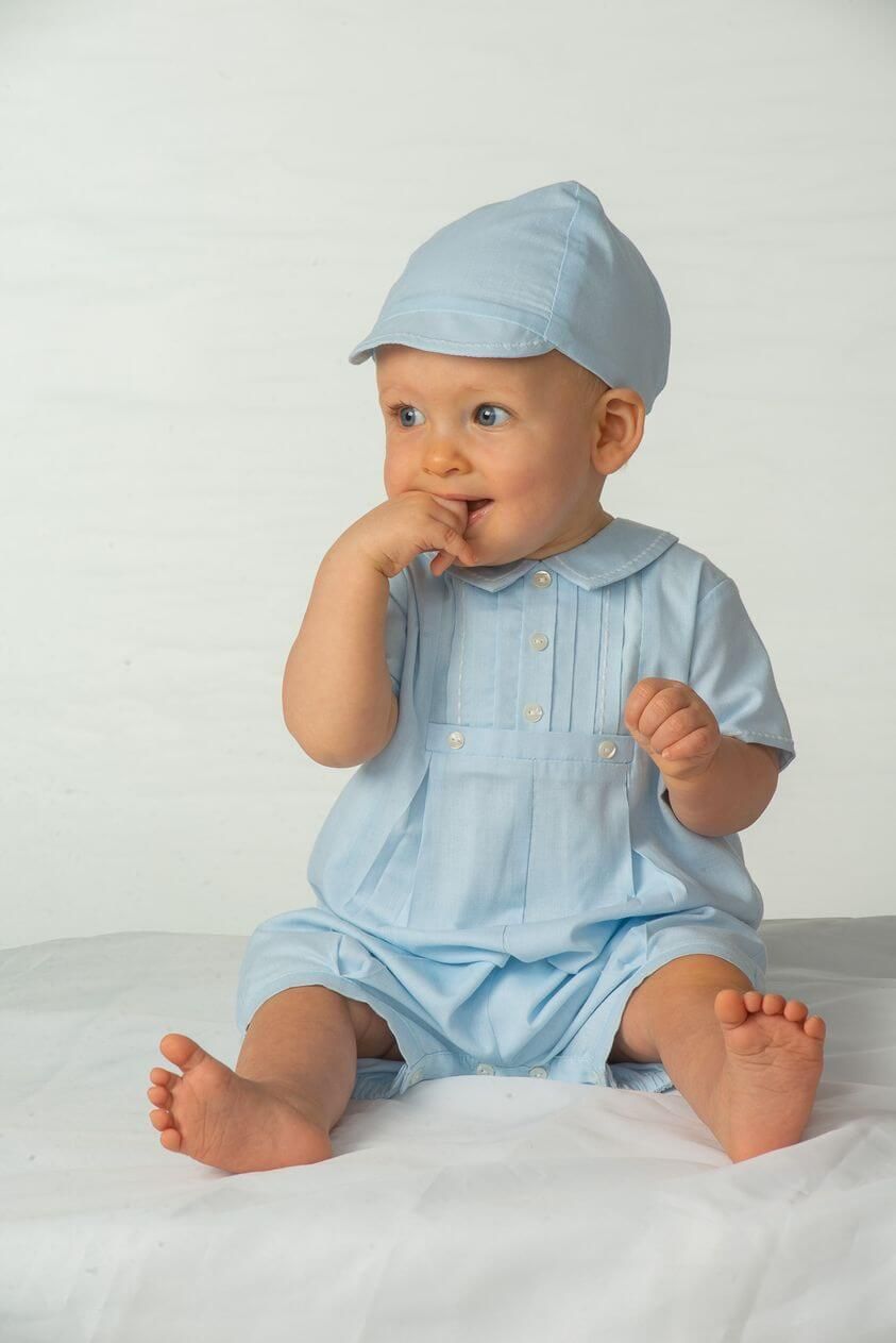 A baby in a blue outfit and hat is sitting on a bed.