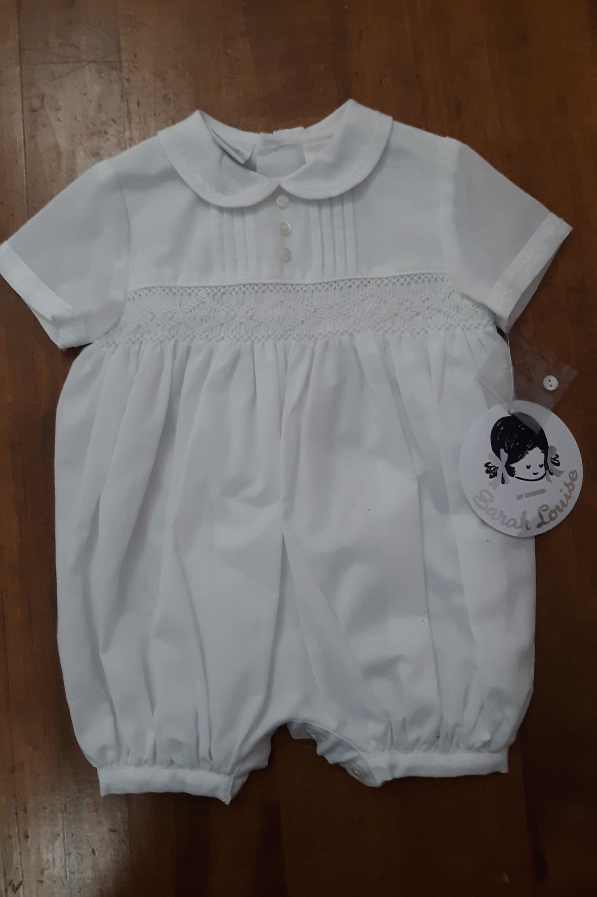 A baby in a white jumpsuit is sitting on a wooden table.