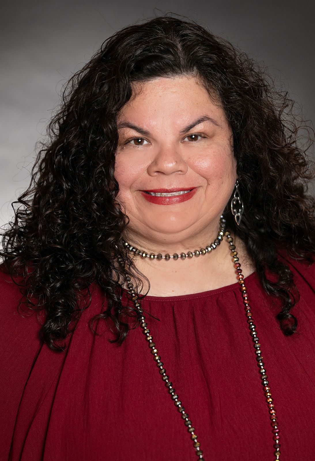 Woman with dark curly hair wearing a maroon top and beaded necklaces, smiling.