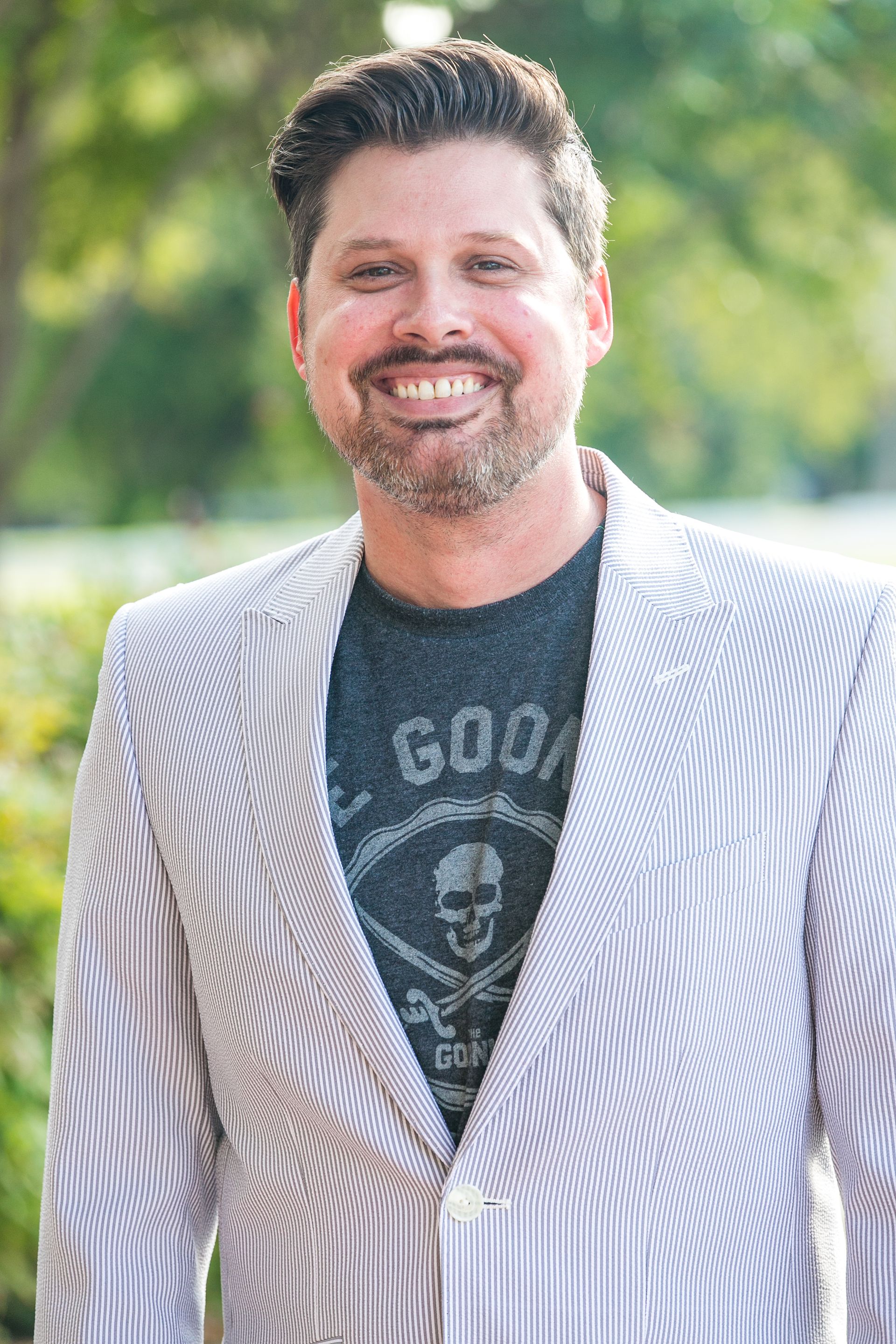Man wearing striped blazer and graphic t-shirt smiles outside.