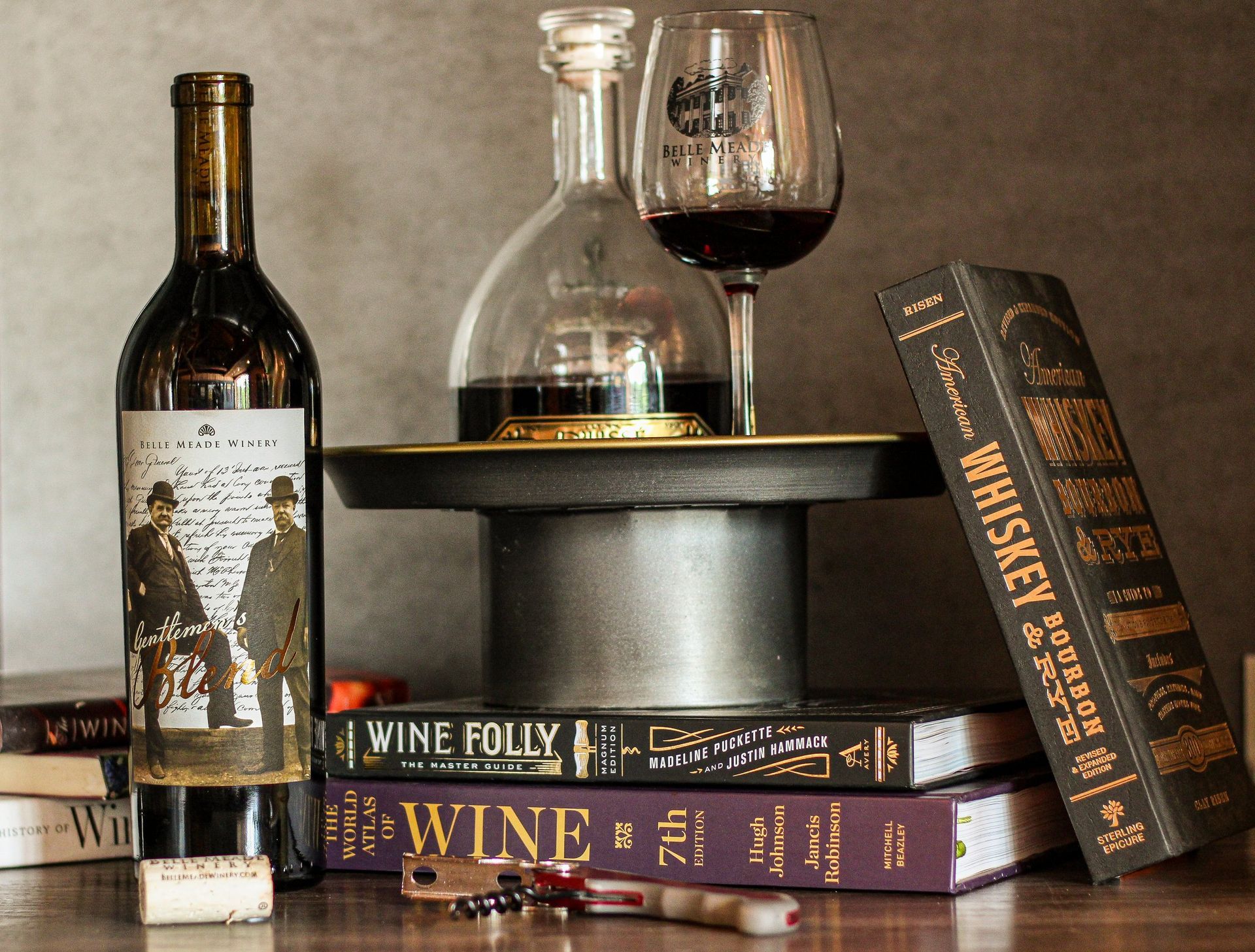 Wine bottle, decanter, glass, books, and corkscrew on a pedestal, with a dark, moody ambiance.
