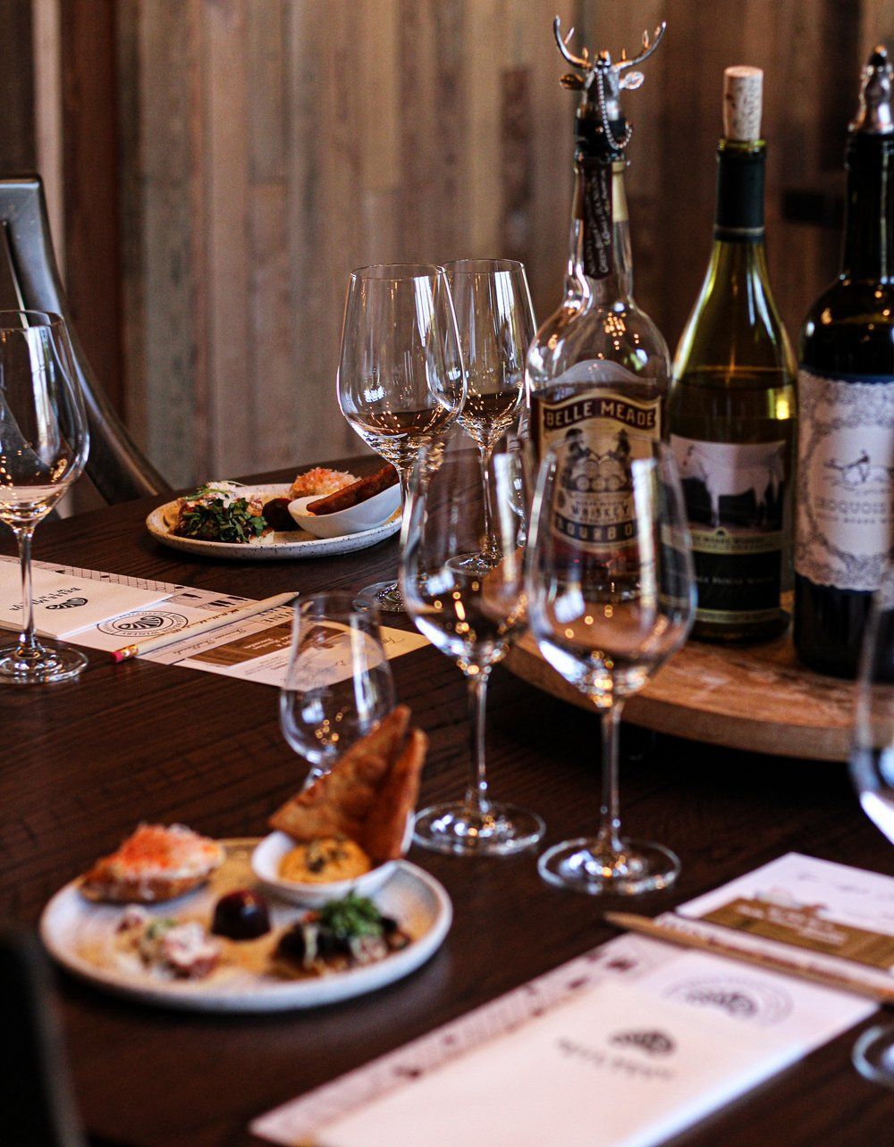 Wine tasting table with glasses, bottles, and small plates of food. Wooden backdrop.