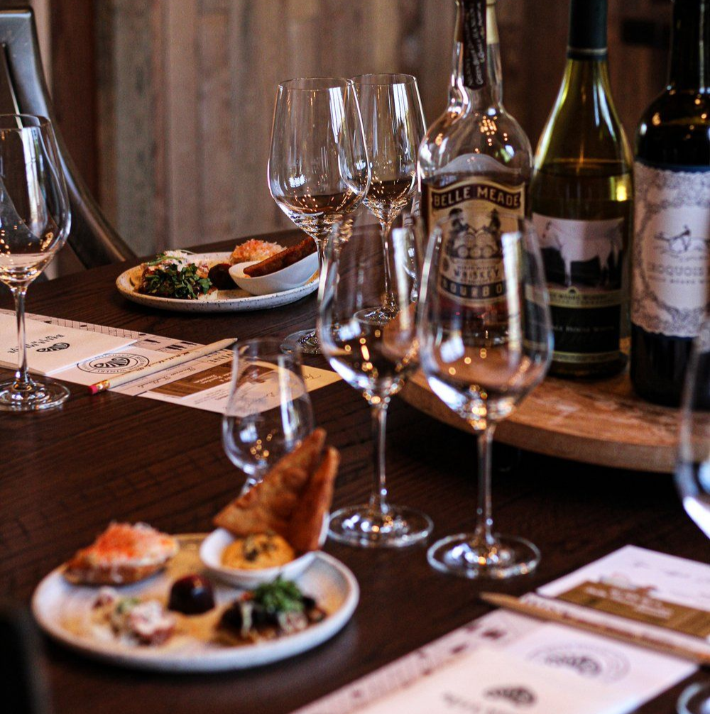 Wine tasting table with glasses, bottles, and small plates of food. Wooden backdrop.