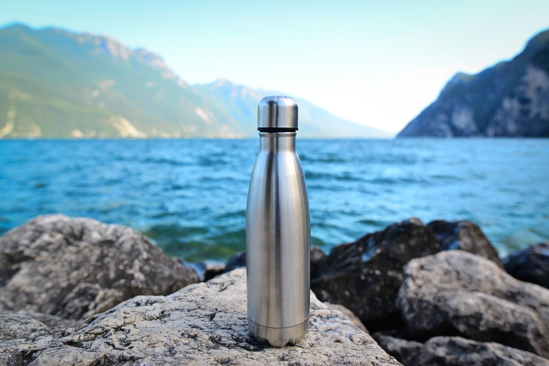 A stainless steel water bottle is sitting on a rock near the water.