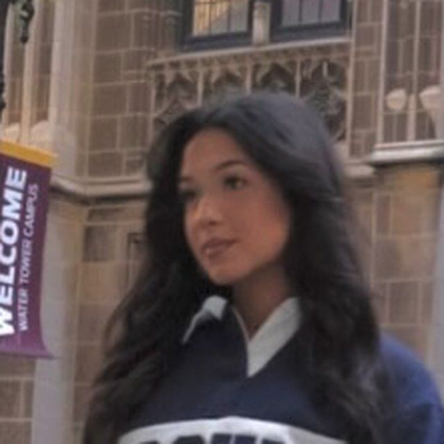 Person with long dark hair in a navy jacket stands outside a stone building with a purple “WELCOME” banner.