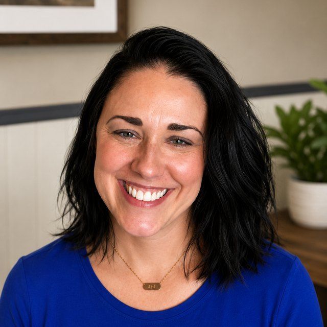 Smiling person in a blue shirt, seated indoors with a plant in the background.