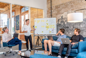 Team meeting in a loft office, with one person presenting at a whiteboard and others seated on a couch.