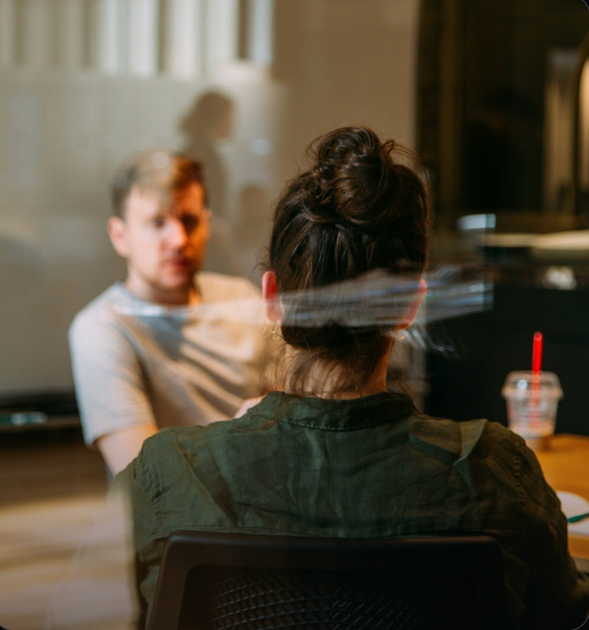 Two people talking across a table in a softly lit cafe, seen through a glass reflection.