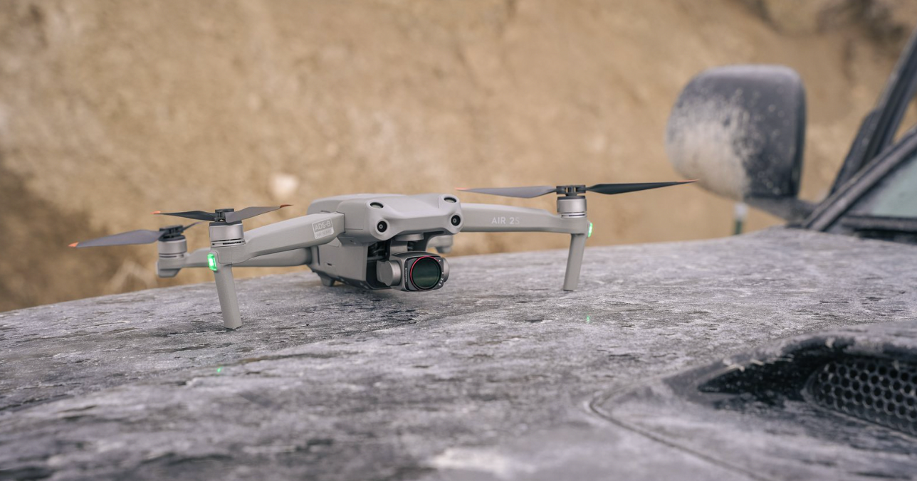 Small gray drone hovering above a dusty car hood in an outdoor setting
