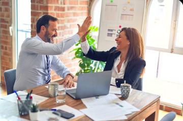 Two coworkers high-five across a desk in a bright office, with a laptop and papers between them.