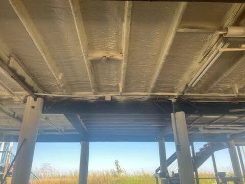 Underside of a raised wooden deck with support posts and beams, overlooking grassy land beyond