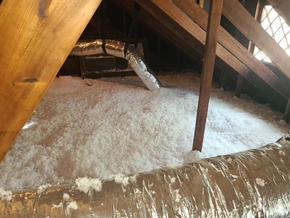 Attic with exposed wooden rafters and a large pile of white insulation around a metal vent pipe