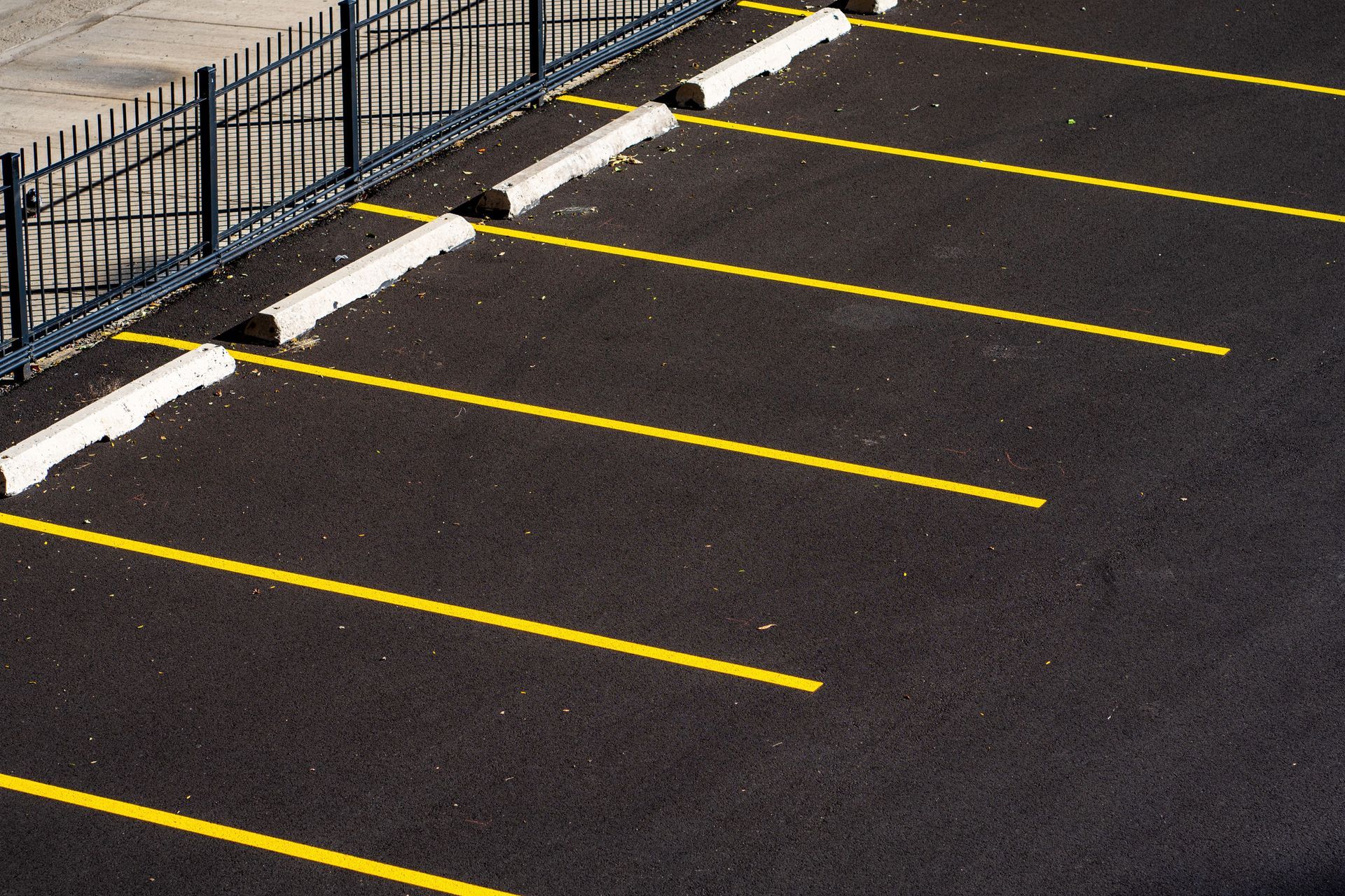 Empty asphalt parking lot with yellow lines beside a black fence and curb stops
