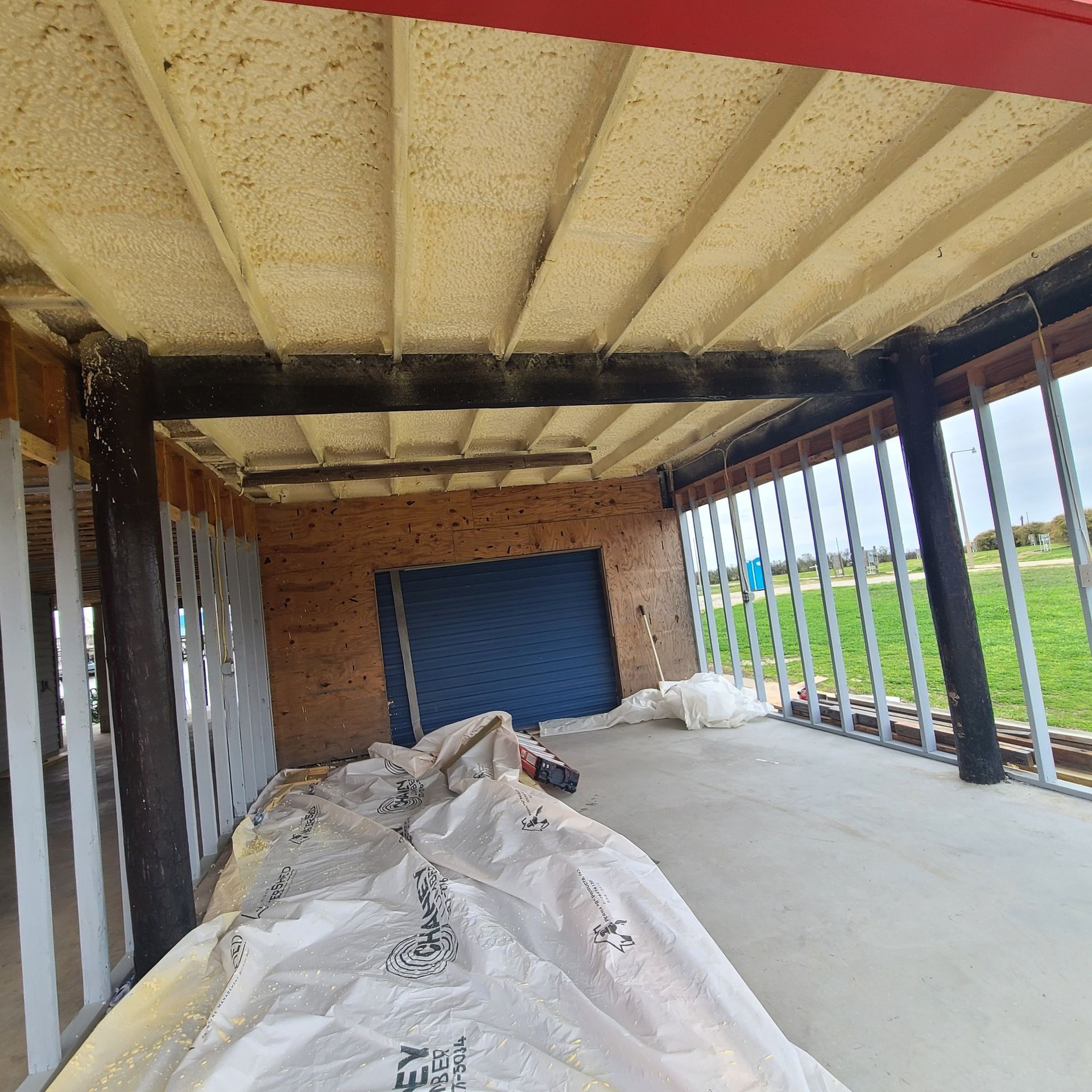 Covered porch under construction with insulation on the ceiling, exposed framing, and a blue door panel at the end.