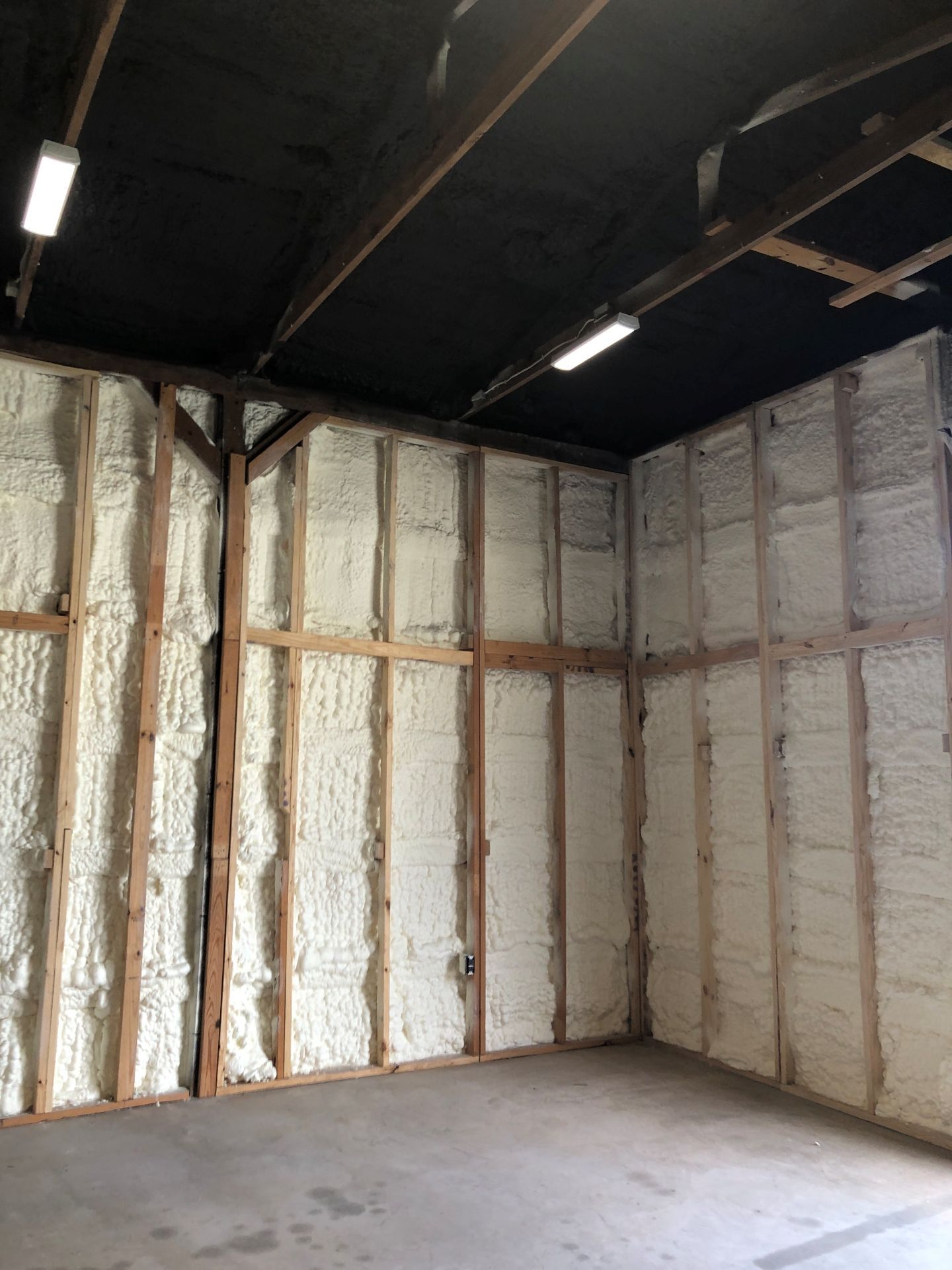 Empty room with exposed insulated walls and a concrete floor, lit by ceiling fluorescent lights.