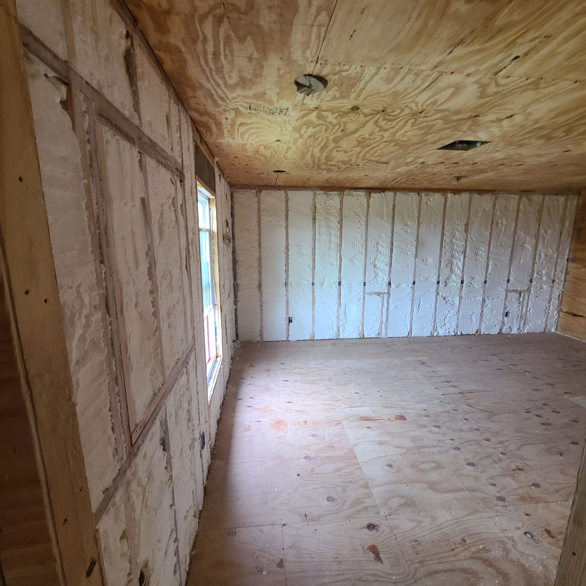 Empty unfinished room with plywood walls, ceiling, and floor, and a window on the left.