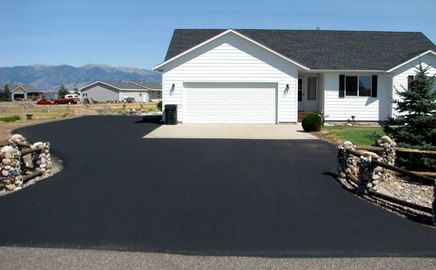 A white house with a black shingled roof features a freshly paved black asphalt driveway flanked by rustic stone fences.