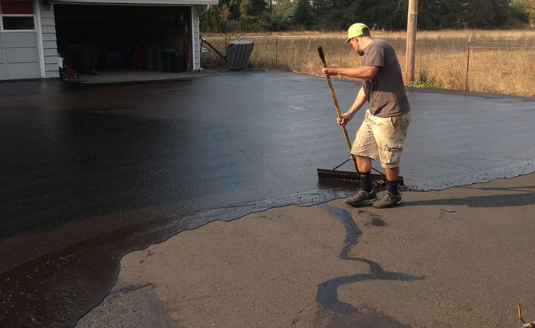A person wearing a neon yellow cap and khaki shorts uses a long-handled brush to spread black sealant on a driveway.