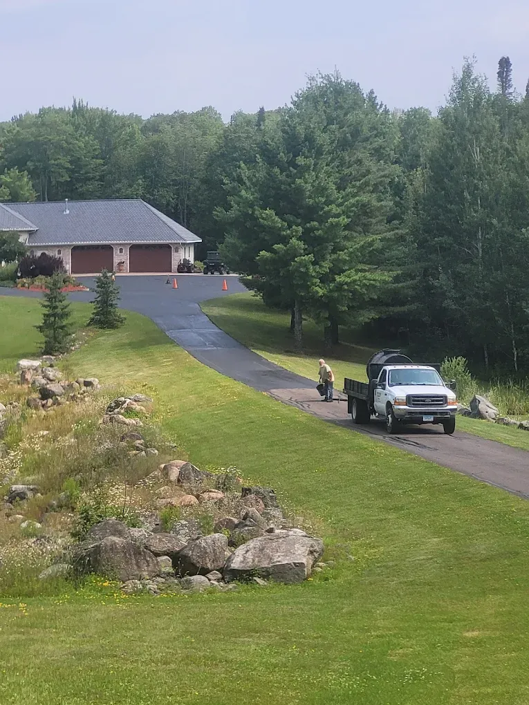 A white pickup truck with a trailer drives up a paved driveway toward a suburban house surrounded by trees.