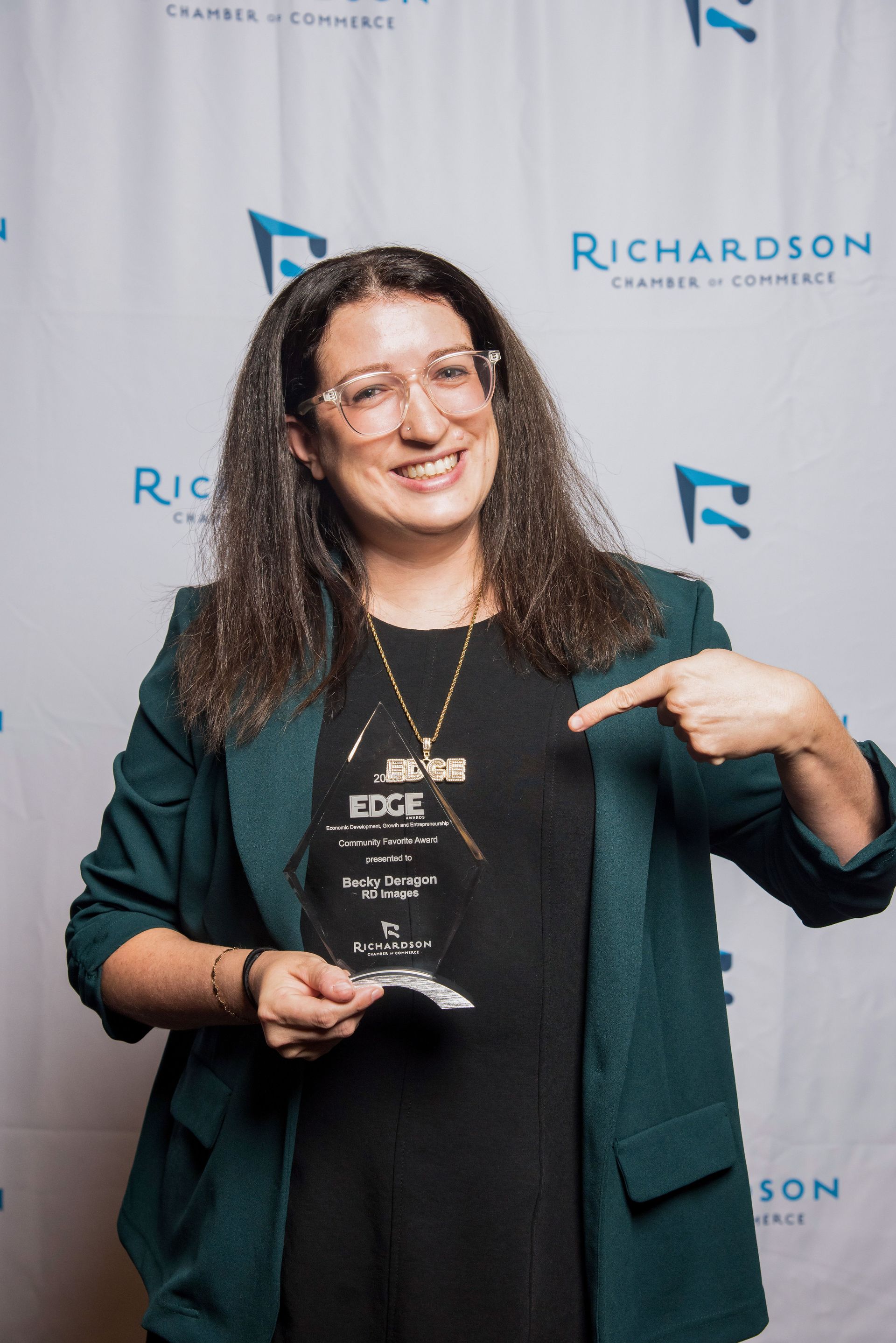 A smiling person in a green blazer holds a glass award in front of a Richardson Chamber of Commerce backdrop.