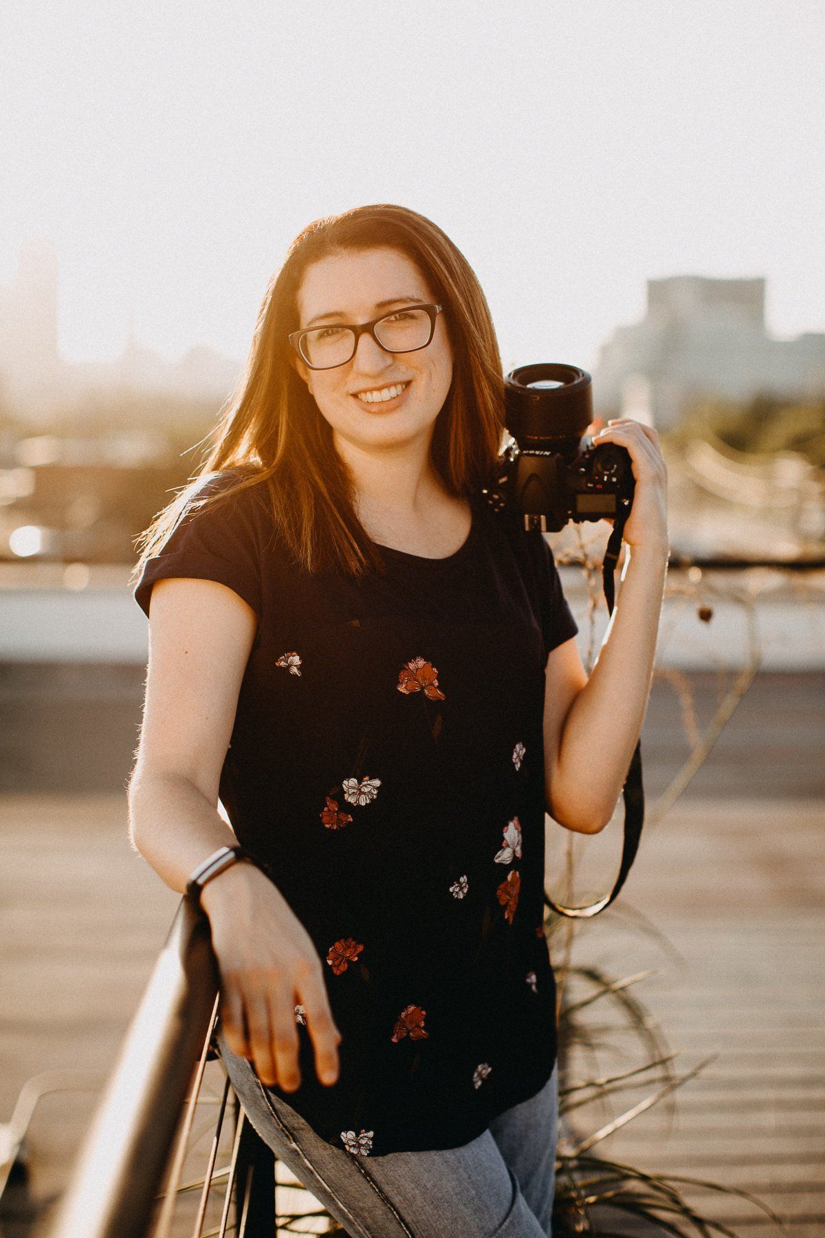 A photographer wearing glasses stands on a balcony at sunset, holding a camera and smiling.