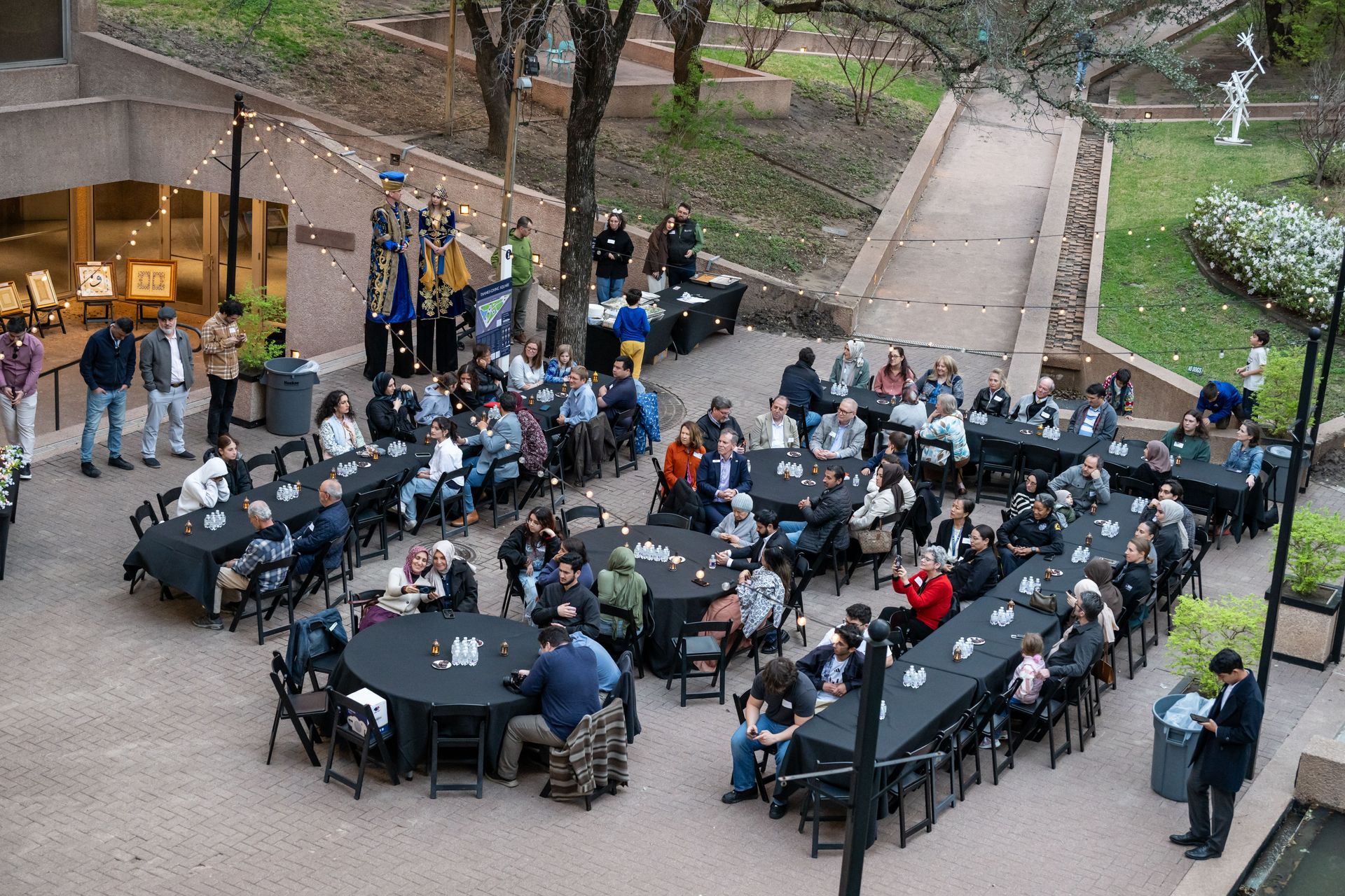 An outdoor evening event features people gathered at round and rectangular tables with black cloths in a courtyard.