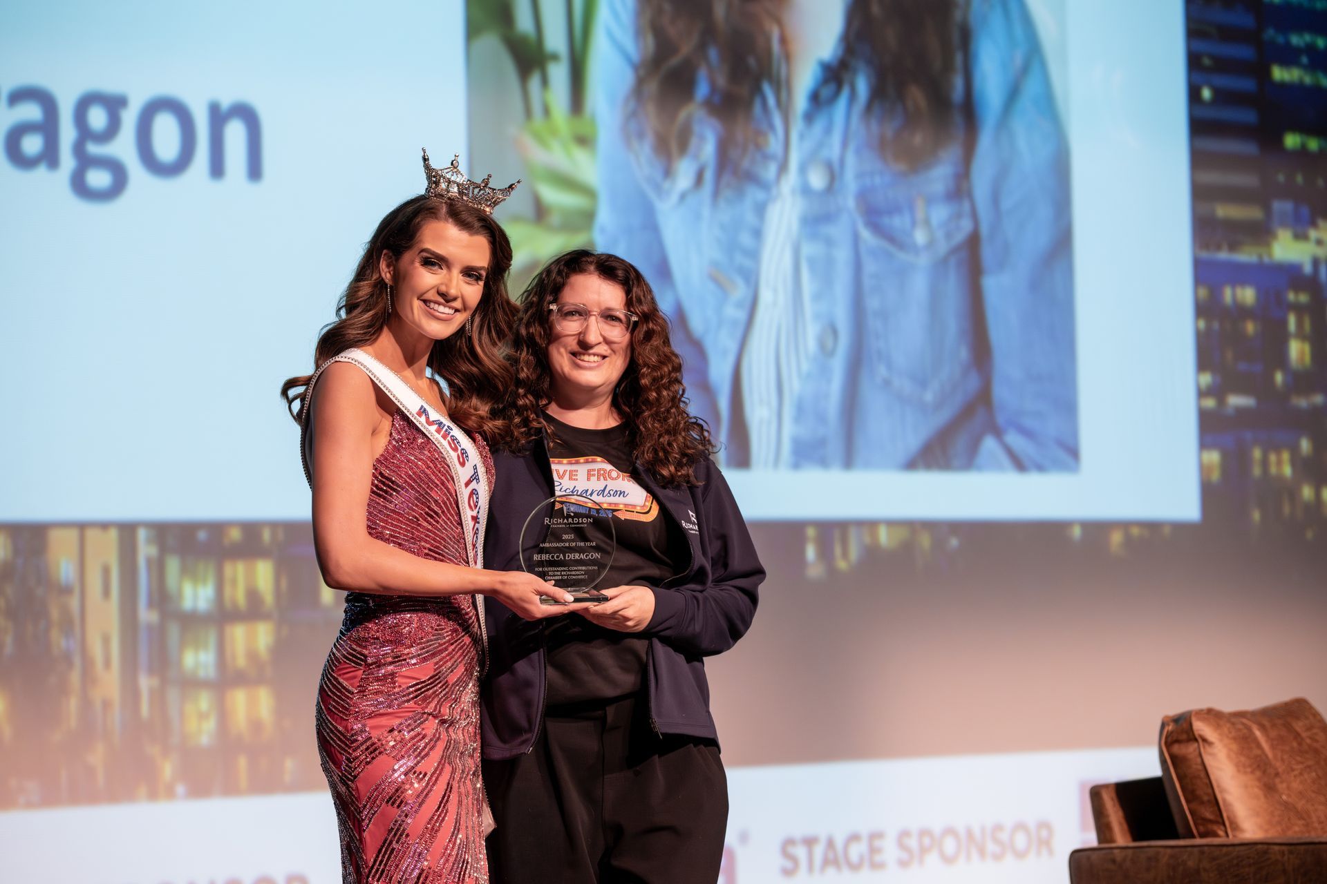 A woman wearing a crown and evening gown stands on stage, handing a small award to another person in casual clothing.
