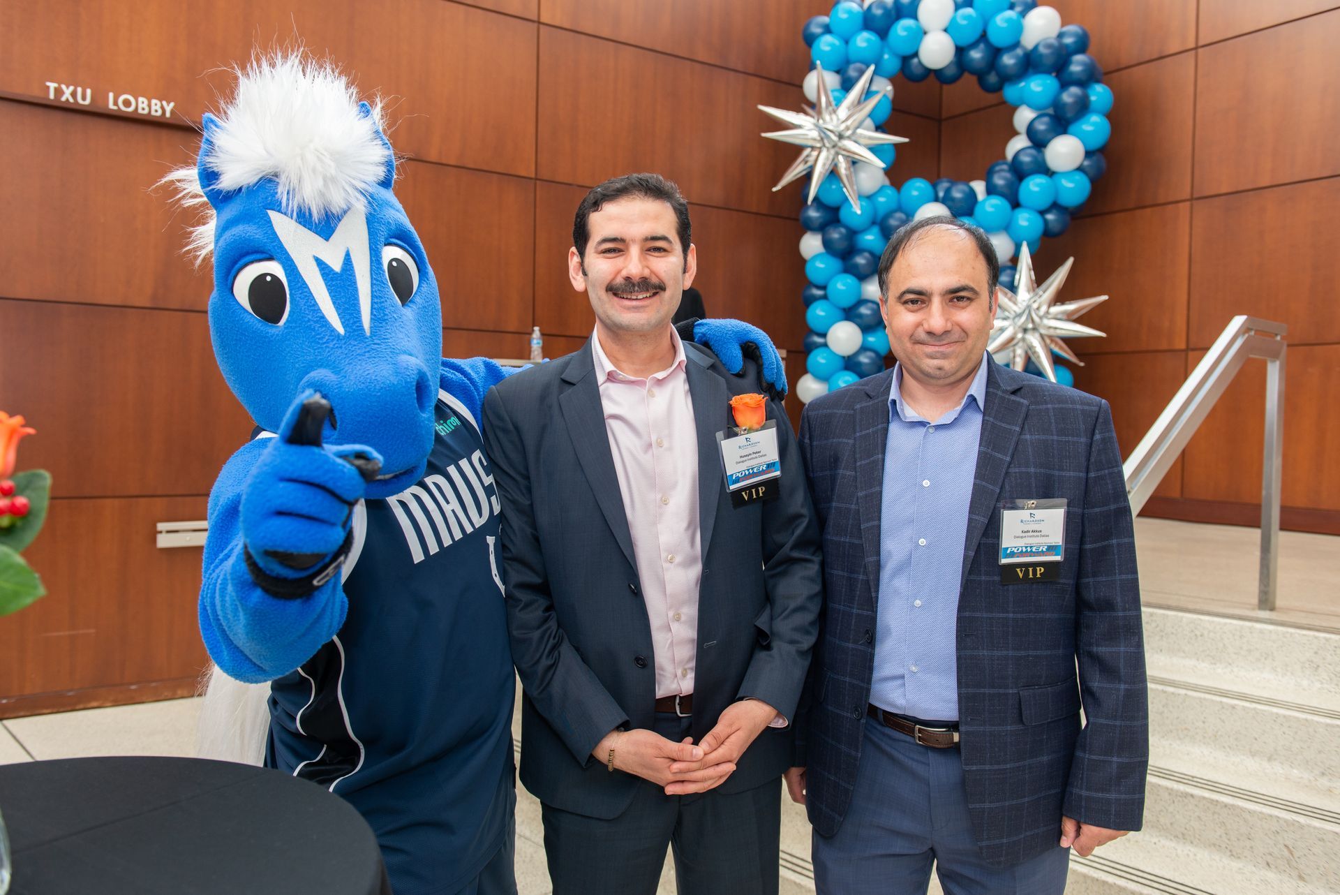 A blue horse mascot poses with two men in suits indoors in front of a blue balloon display and wooden wall panels.