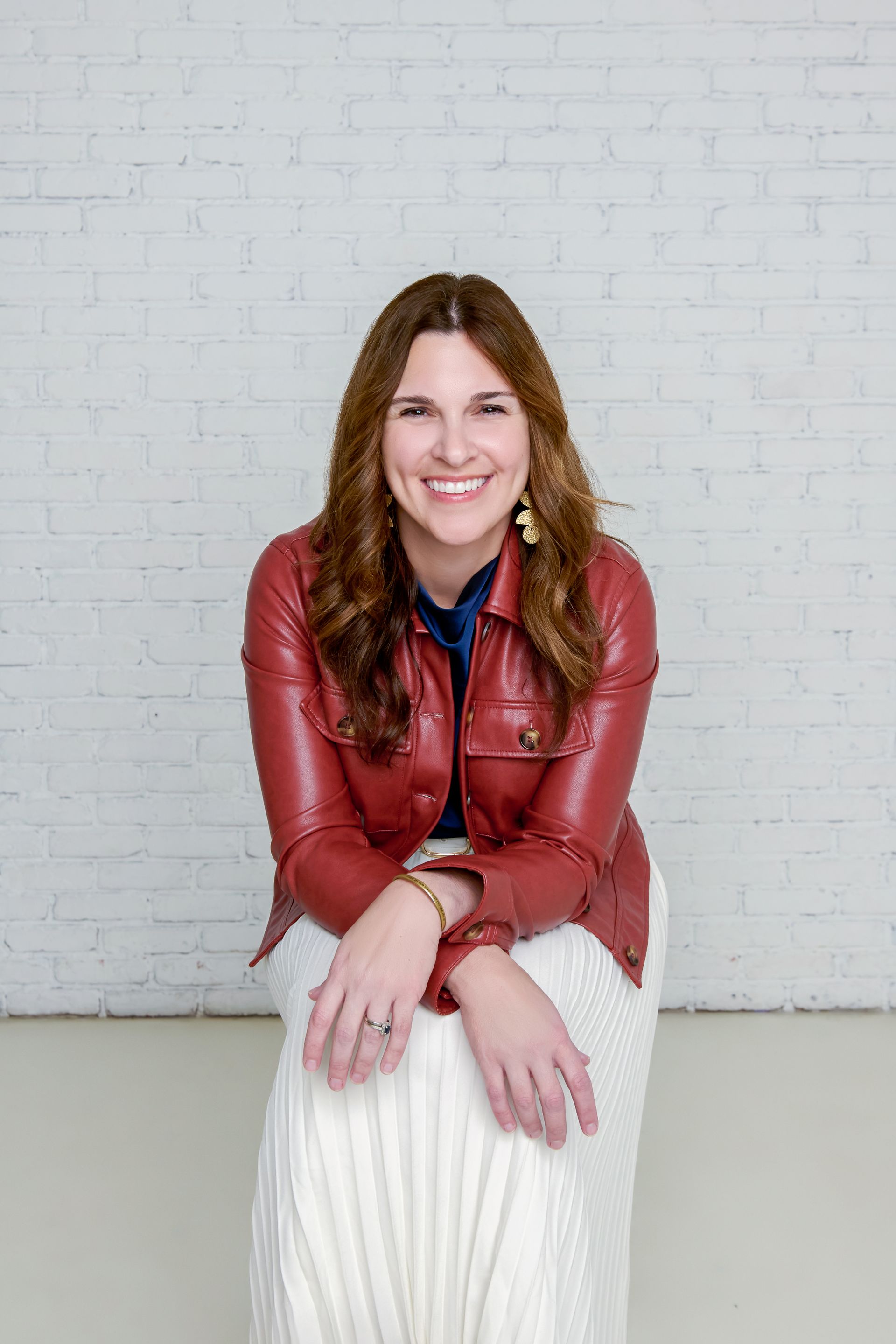 A smiling person wears a red leather jacket and a long, white, textured skirt while sitting against a white brick wall.