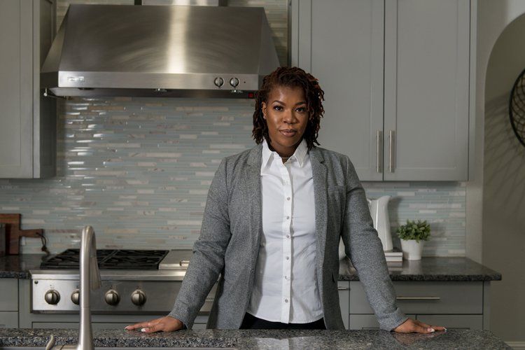 A person in a professional grey blazer and white shirt stands behind a kitchen counter in a modern, neutral-toned kitchen.