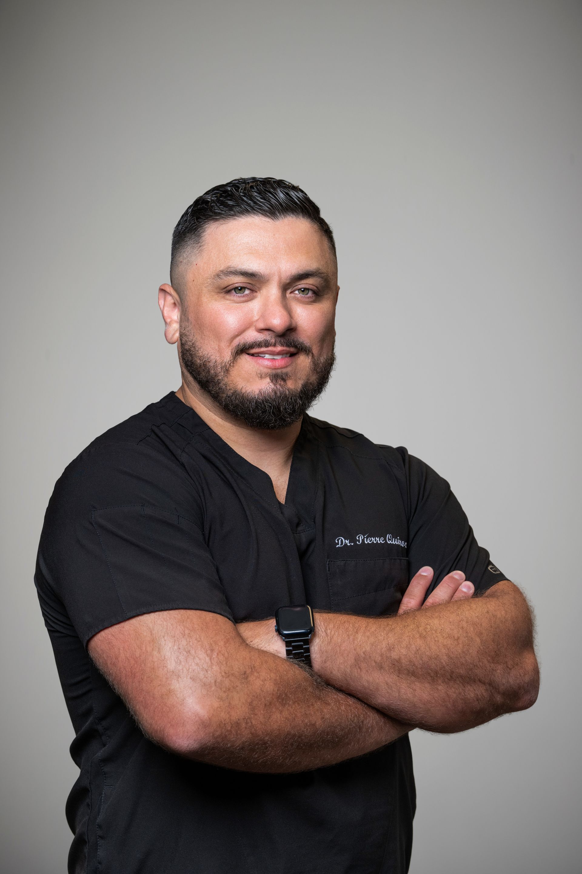 A person with short dark hair and a beard, wearing a black scrub top with arms crossed, posing against a light gray background.
