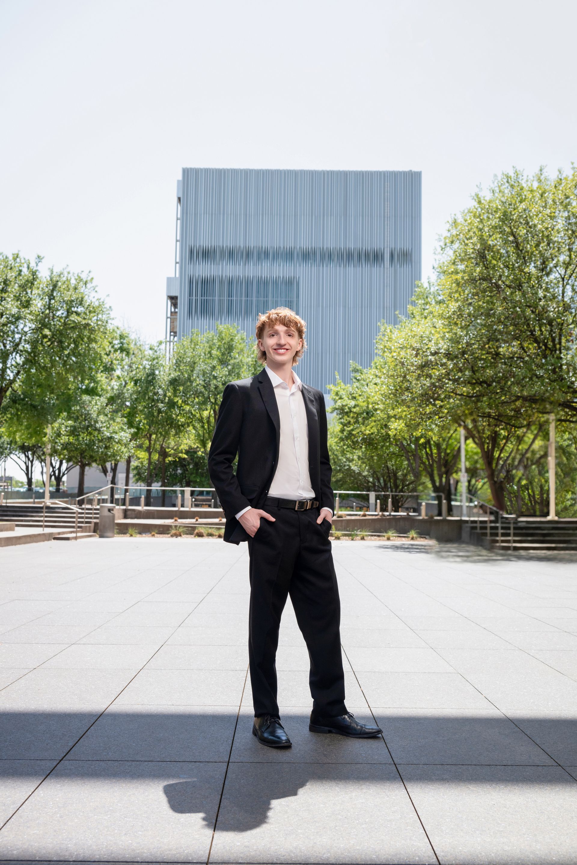 A person in a black suit and white shirt standing on a paved plaza with trees and a modern building in the background.