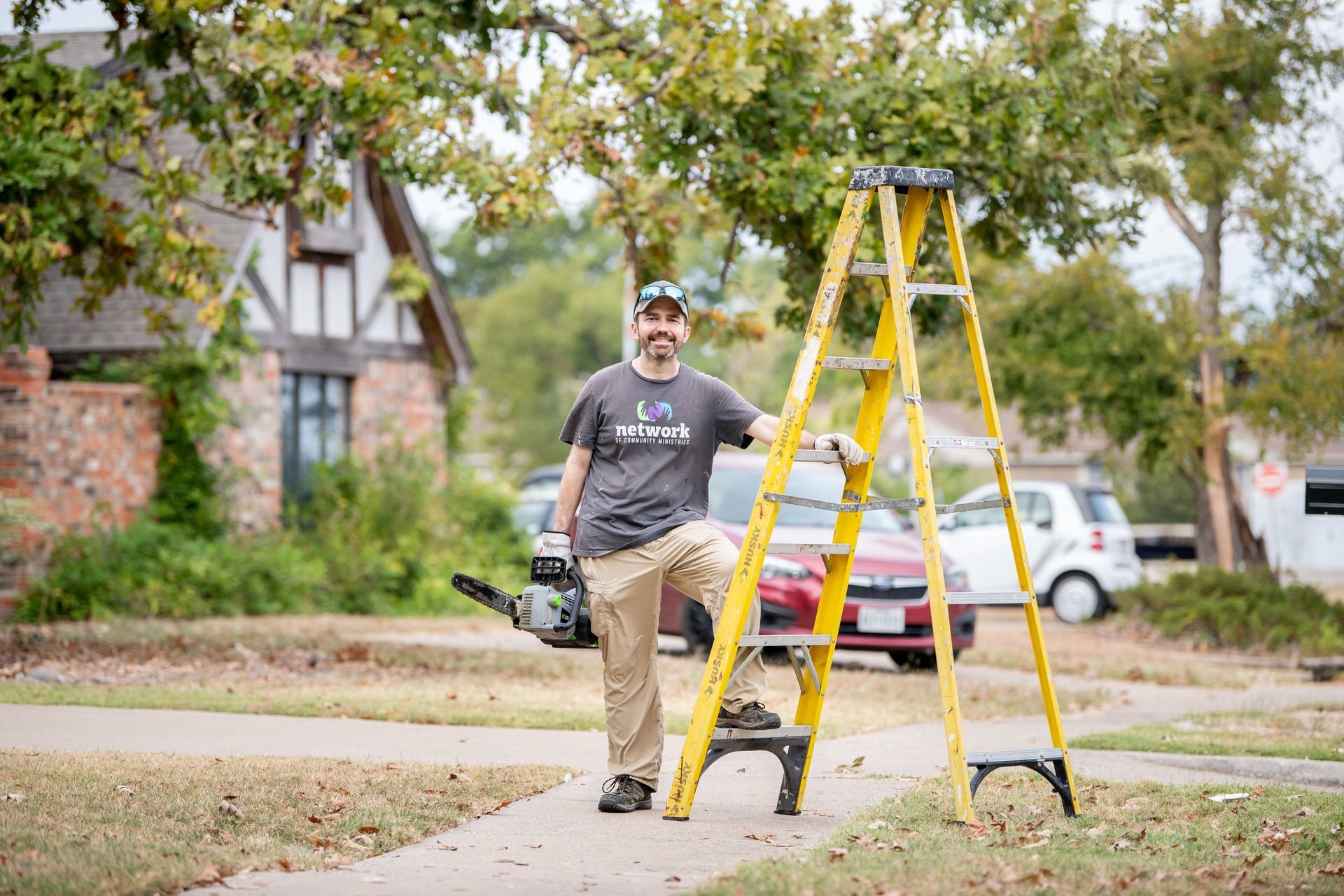 A person wearing a grey shirt and khakis stands on a walkway next to a yellow ladder, holding a chainsaw.