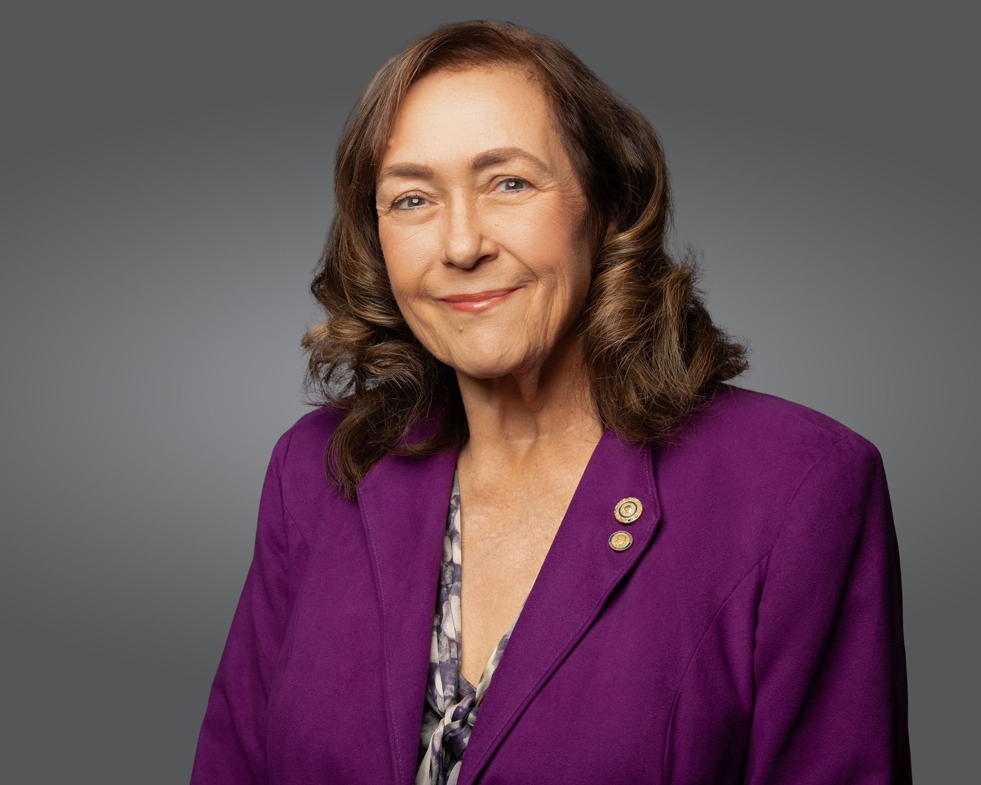 A portrait of a person with shoulder-length brown hair, wearing a vibrant purple blazer over a patterned top.