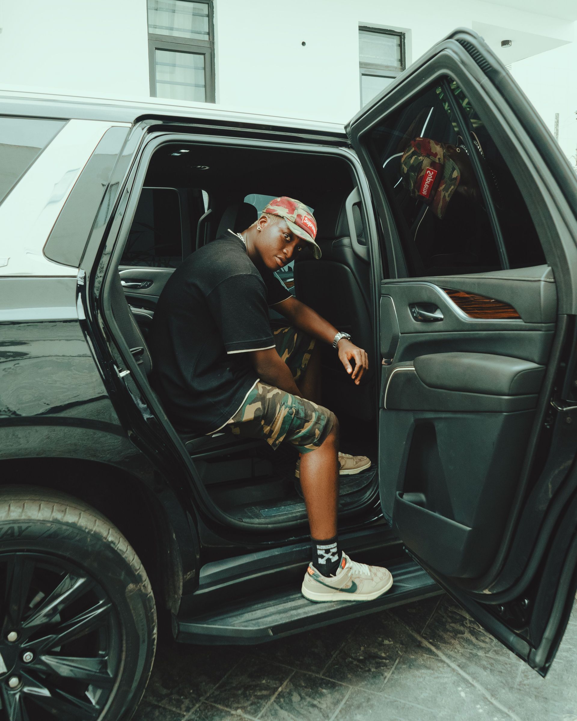 Person sitting in the open rear door of a black SUV, wearing a cap and shorts.