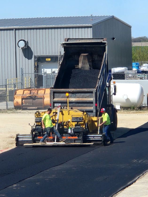 Workers operate a road machine beside a dump truck in an industrial lot.